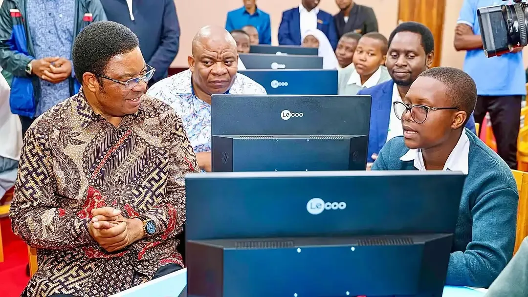 A student demonstrates work on a desktop computer to a former prime minister Kassimu Majaliwa in a school computer lab, while other officials and students watch from rows of computers in the background.