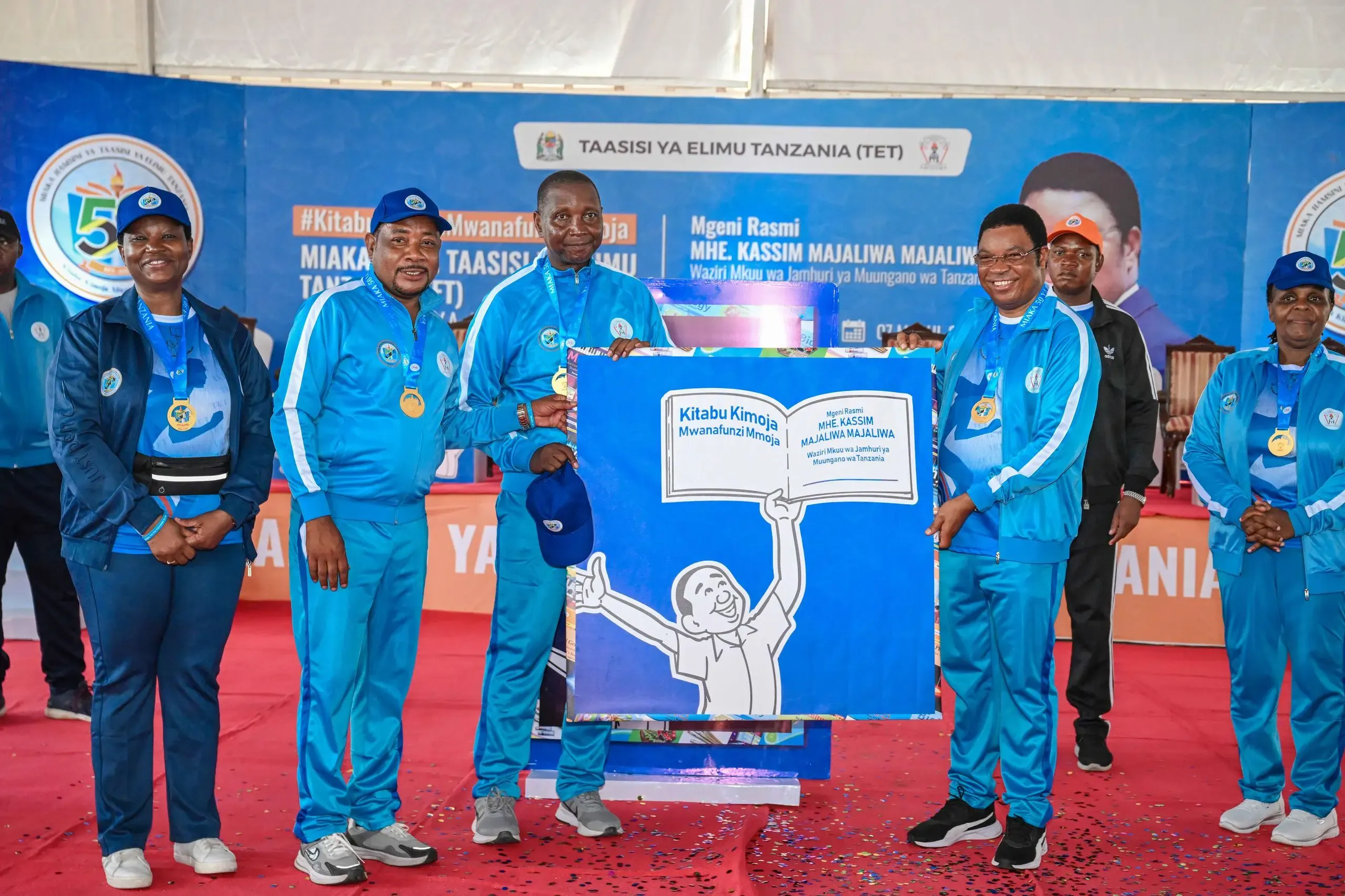 Minister of Education Adolf Mkenda, the Director General of the Tanzania Institute of Education Anneth Komba, a former prime minister kassim majaliwa, and other officials stand on a stage holding a large ‘Kitabu kimoja mwanafunzi mmoja campaign display during the launch event celebrating 50 years of the Tanzania Institute of Education.