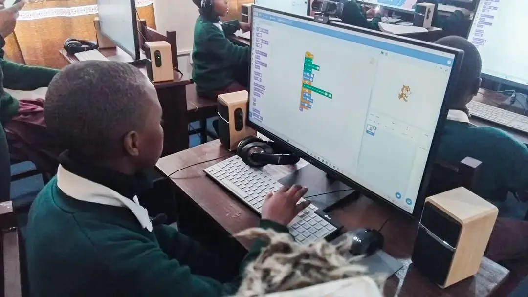 Student in a computer lab uses a desktop computer to build a Scratch-style coding program, with other students working at nearby machines in the background.