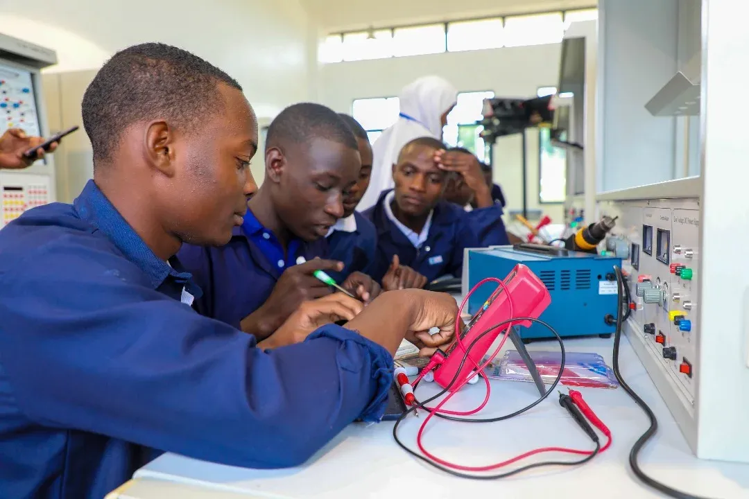 Students in blue uniforms work together at a science lab bench, using a multimeter and wires to test an electronics setup while classmates watch and take notes.