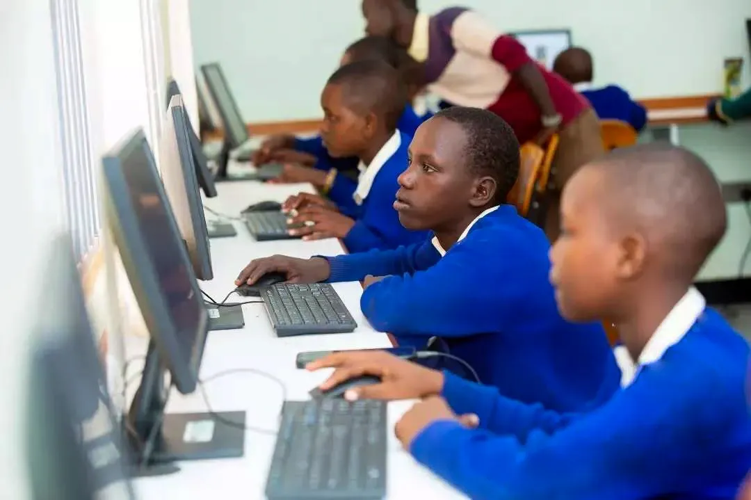 Students in blue school uniforms sit in a computer lab, working at desktop computers in a row while a teacher assists in the background.