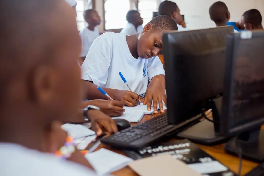 Student in a white T-shirt writes in a notebook at a computer desk, with desktop monitors and other students working in the background.