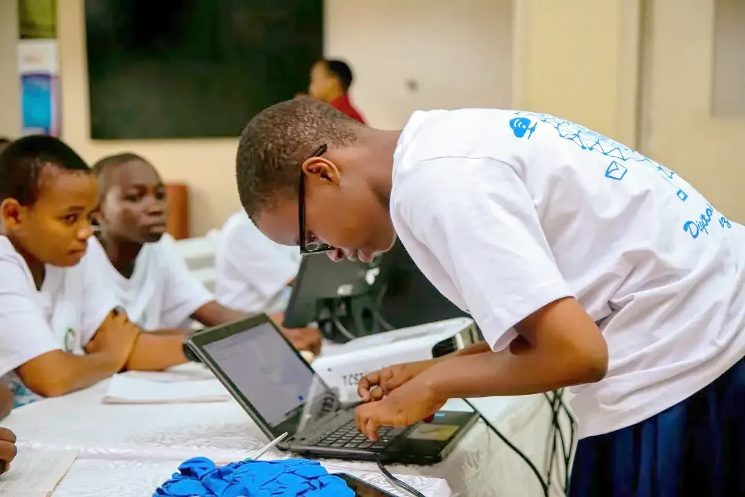 A female student in a white T-shirt leans over a laptop, typing or adjusting settings, while other students sit at the same table watching in a classroom.