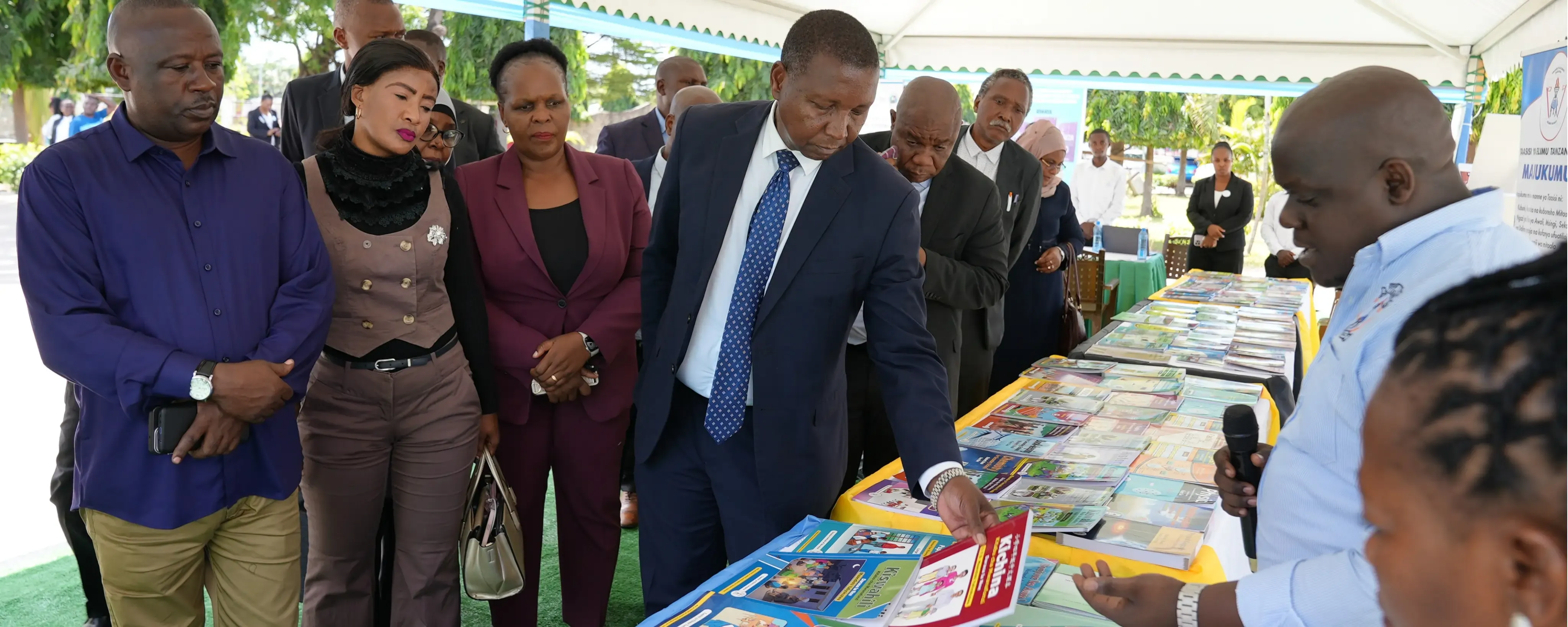 Minister of Education Adolf Mkenda and the Director General of the Tanzania Institute of Education Anneth Komba stand with other officials under a tent, looking at textbooks and learning materials displayed on tables while a presenter explains them.